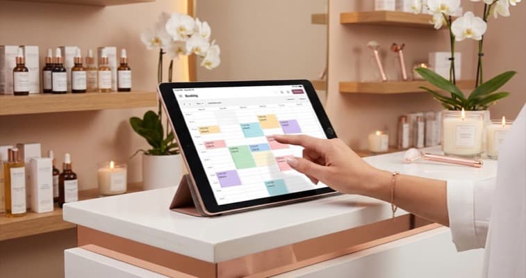 A beauty therapist touches a tablet screen showing a booking interface on a salon reception counter with treatment products in the background