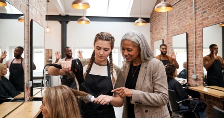 Young hairdressing apprentice learning to style hair in a modern UK salon supervised by a senior stylist