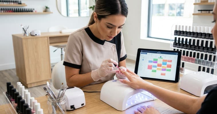 Nail technician working on gel nails with a nail salon booking system shown on a tablet beside the UV lamp