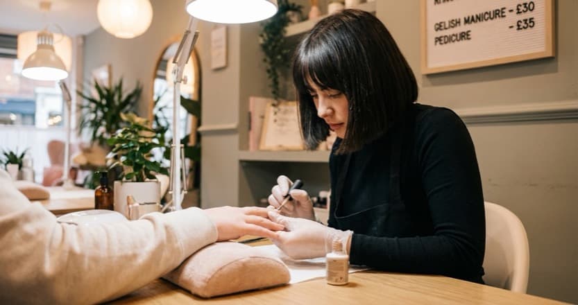 Nail technician applying gel polish at a nail desk with a printed pricing board visible in the background